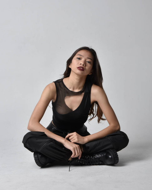 Full length portrait of pretty young asian girl wearing black tank top, utilitarian  pants and leather boots. Sitting poseisolated against a  studio background. - Photo, image