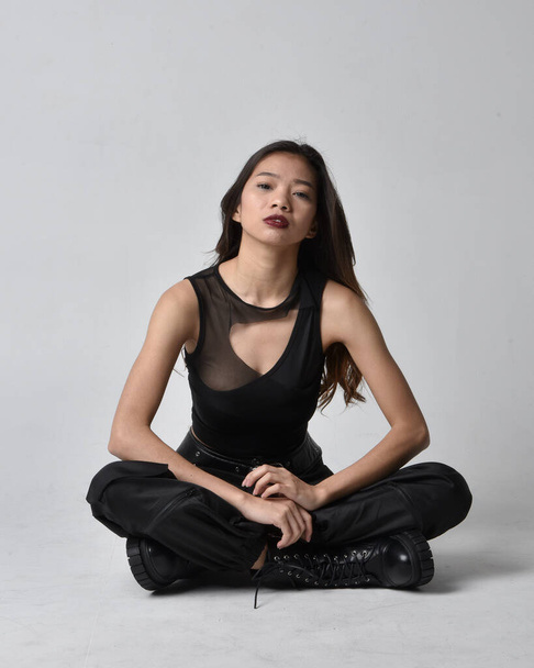 Full length portret van mooie jonge aziatische meisje dragen zwarte tank top, utilitaire broek en lederen laarzen. Zittende houding geïsoleerd tegen een studio achtergrond. - Foto, afbeelding