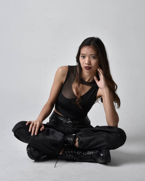 Portret młodej Azjatki w czarnej koszulce, użytkowych spodniach i skórzanych butach. Siedzenie w izolatce na tle studia. - Zdjęcie, obraz