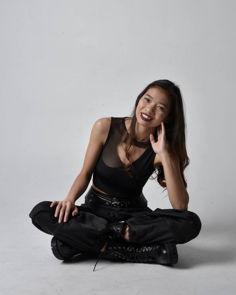 Full length portret van mooie jonge aziatische meisje dragen zwarte tank top, utilitaire broek en lederen laarzen. Zittende houding geïsoleerd tegen een studio achtergrond. - Foto, afbeelding
