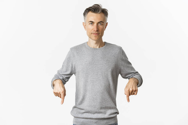 Portrait d'un homme d'âge moyen en pull gris, pointant les doigts vers le bas et regardant avec confiance la caméra, montrant le logo, debout sur fond blanc - Photo, image