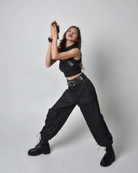 Ganzkörperporträt eines hübschen jungen asiatischen Mädchens mit schwarzem Tank-Top, praktischer Hose und Lederstiefeln. Stehende Pose mit Waffe, isoliert vor Studiohintergrund. - Foto, Bild