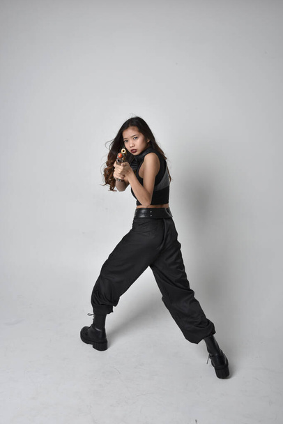 Full length portret van mooie jonge aziatische meisje dragen zwarte tank top, utilitaire broek en lederen laarzen. Staande houding met een pistool, geïsoleerd tegen een studio achtergrond. - Foto, afbeelding