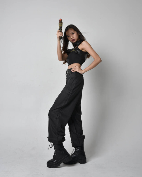 Full length portret van mooie jonge aziatische meisje dragen zwarte tank top, utilitaire broek en lederen laarzen. Staande houding met een pistool, geïsoleerd tegen een studio achtergrond. - Foto, afbeelding