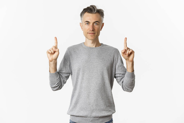 Porträt eines gutaussehenden Mannes mittleren Alters in grauem Pullover, der Promo zeigt, mit erhobenem Finger ernst guckt und vor weißem Hintergrund steht - Foto, Bild
