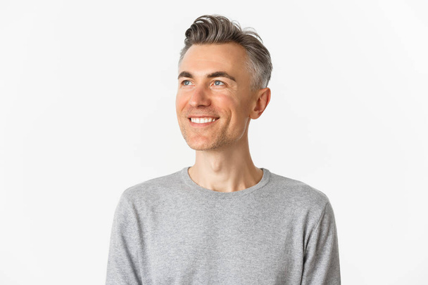 Close-up van tevreden man van middelbare leeftijd, glimlachend en kijkend naar linksboven, staand in grijze trui over witte achtergrond - Foto, afbeelding