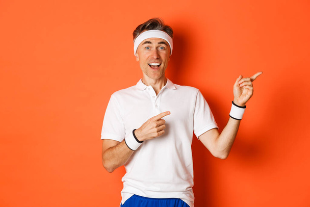 Koncept cvičení, sportu a životního stylu. Portrét pohledného muže středního věku ve sportovním oblečení, ukazuje prsty v pravém horním rohu a vypadá ohromeně, ukazuje logo, oranžové pozadí - Fotografie, Obrázek