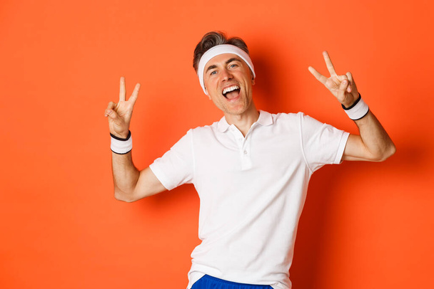 Immagine di felice e sano uomo di mezza età in abbigliamento sportivo, mostrando segni di pace e sorridente, allenamento su sfondo arancione - Foto, immagini