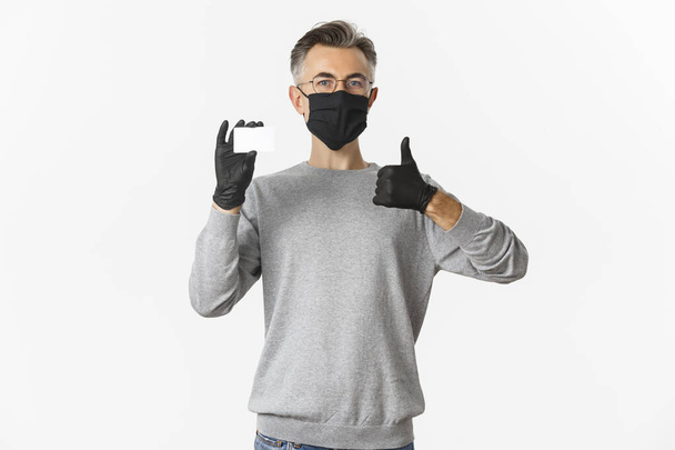 Concept van covid-19, sociale afstand en levensstijl. Tevreden man van middelbare leeftijd met medisch masker, handschoenen en bril, aanbevolen bank, met kredietkaart en duim omhoog - Foto, afbeelding