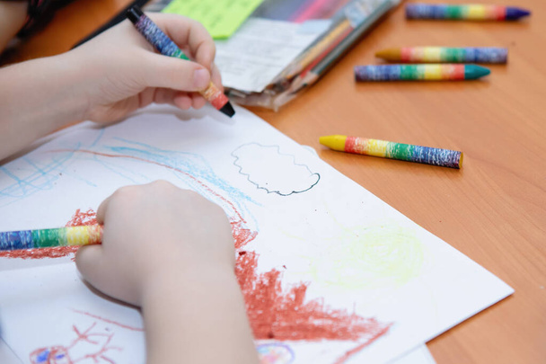 Dětské ruce kreslí voskovou křídou na bílém listu papíru na stole. Školní základní vzdělání. Selektivní soustředění. Rozmazané pozadí. - Fotografie, Obrázek