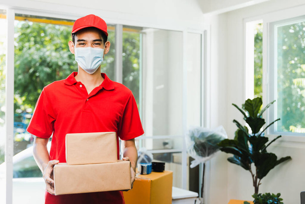 Profesjonalny azjatycki doręczyciel w czerwonym mundurze noszący maskę twarzy trzymającą skrzynkę ładunkową i paczkę w domu, usługę przechowywania w domu i koncepcję wysyłki - Zdjęcie, obraz