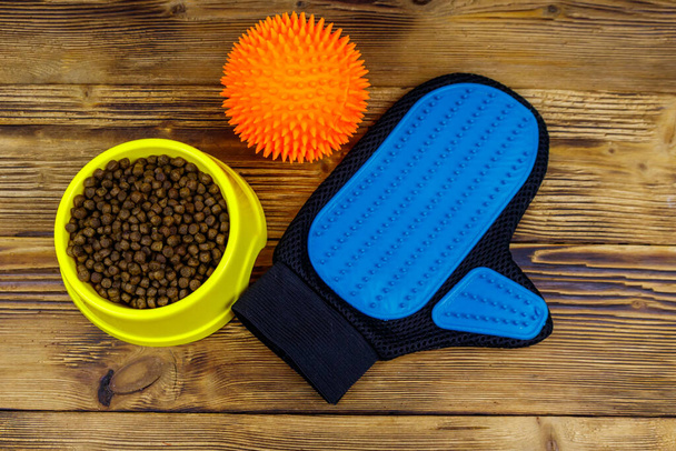 Dry dog food in bowl, ball toy and pet grooming glove on wooden background. Top view. Pet care concept - Photo, Image