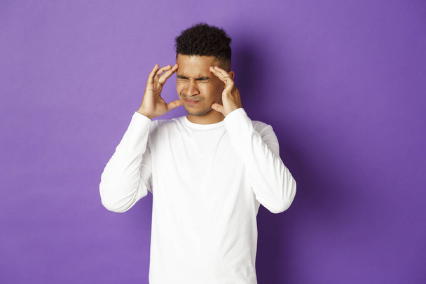 Изображение красивого африканско-американского мужчины, страдающего головной болью, гримасой и трогательной головой, страдающего мигренью, стоящего на фиолетовом фоне - Фото, изображение