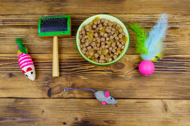 Konzervované kočičí jídlo v misce, kočičí hračky a mazlíček slicker kartáč na dřevěném pozadí. Horní pohled. Koncept péče o zvířata - Fotografie, Obrázek