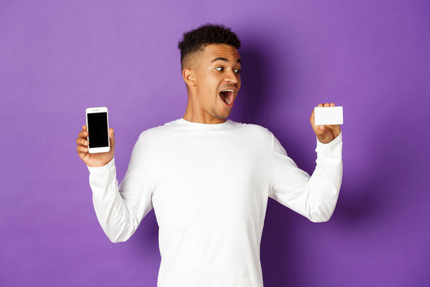 Portrait of handsome african-american man, looking amazed at credit card and showing app on mobile phone screen, standing over purple background - Photo, Image