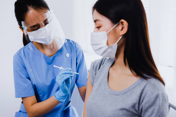 Fiatal ázsiai nő ül az orvosi rendelőben és kap egy influenza oltást a karjába. Orvosi arcmaszkban lévő nővér, aki fecskendőt tart és modern Covid 19 vakcinát ad be a női betegnek - Fotó, kép