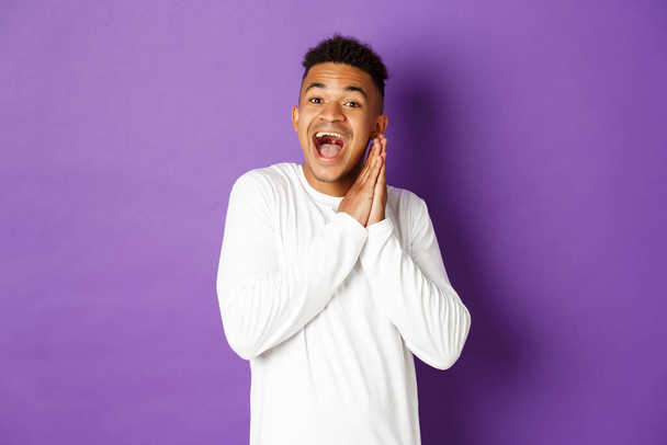 Afbeelding van opgewonden en hoopvolle Afrikaans-Amerikaanse homo, op zoek met spanning en sensatie naar iets, klappende handen en glimlachend gelukkig, staande over paarse achtergrond - Foto, afbeelding