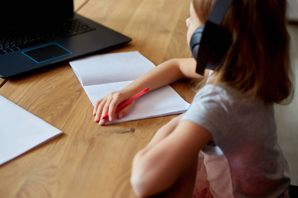 Симпатичная маленькая девочка с наушниками, используя ноутбук, чтобы учиться дома, писать, отвечать, онлайн-обучение, образование, социальная дистанция, карантин коронавируса. - Фото, изображение