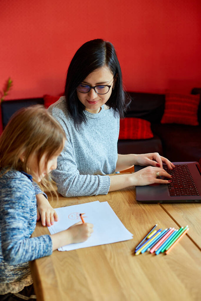 Moeder werken in haar kantoor thuis op een laptop, haar dochter zit naast haar en tekenen, Vrouw freelance, afgelegen werk en het opvoeden van een kind op het werk. - Foto, afbeelding