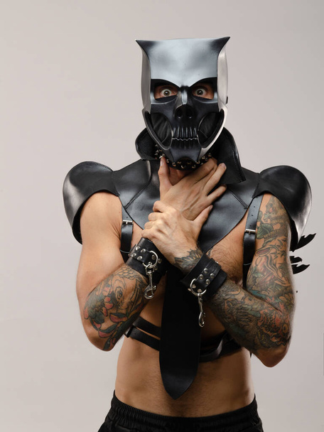 un hombre con una máscara de cráneo demonio bdsm, vestido con una capa de cuero con pulseras de cuero y correas en su cuerpo - Foto, imagen