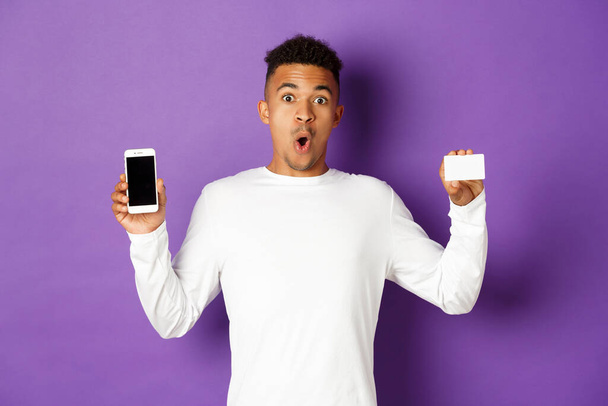 Portret przystojnego, afrykańsko-amerykańskiego mężczyzny, wyglądającego na zdumionego, pokazującego ekran smartfona i kartę kredytową, stojącego na fioletowym tle - Zdjęcie, obraz