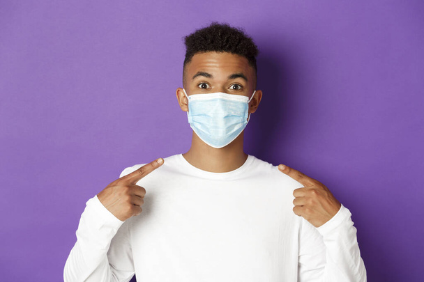 Pojęcie koronawirusu, kwarantanny i dystansu społecznego. Zbliżenie młodego afro-amerykanina uśmiechającego się, polecającego noszenie maski medycznej podczas covid-19, stojącego nad fioletowym tłem - Zdjęcie, obraz