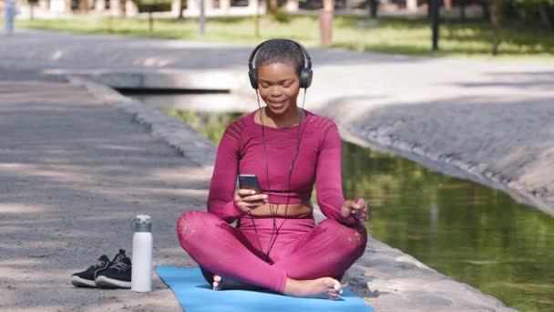 Portret van positieve plus size goed passende dame in sportkleding ontspannen op yoga mat buiten, glimlachen mooie vrouwelijke fitnesstrainer luisteren muziek met oortelefoons alleen. Sport actief lifestyle concept - Video