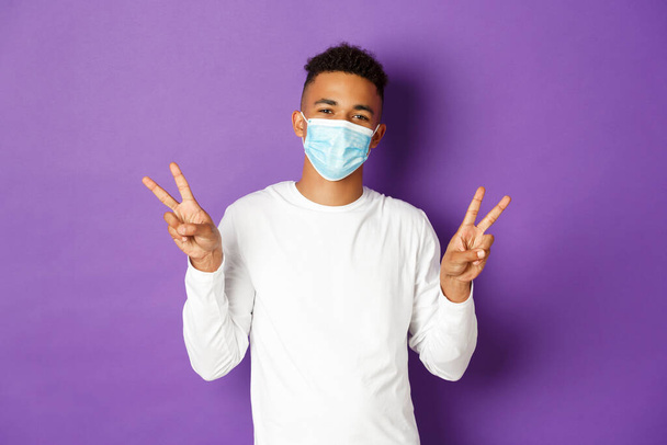 Concetto di covid-19, pandemia e distanza sociale. Carino ragazzo afro-americano in maschera medica, sorridente e mostrando segni di pace, in piedi su sfondo viola - Foto, immagini