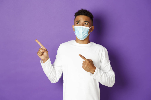 Koncepcja koronawirusu, kwarantanny i stylu życia. Zszokowany afro-amerykański mężczyzna, noszący maskę medyczną, wskazujący i wyglądający nerwowo w lewym górnym rogu, stojący nad fioletowym tłem - Zdjęcie, obraz