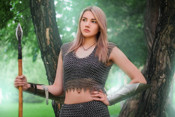 Portrait of a young medieval woman warrior, dressed in chain mail armor with a spear in her hands against the background of the dark forest. - Foto, Bild