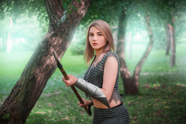 Portrait of a young medieval woman warrior, dressed in chain mail armor with a spear in her hands against the background of the dark forest. - Фото, зображення