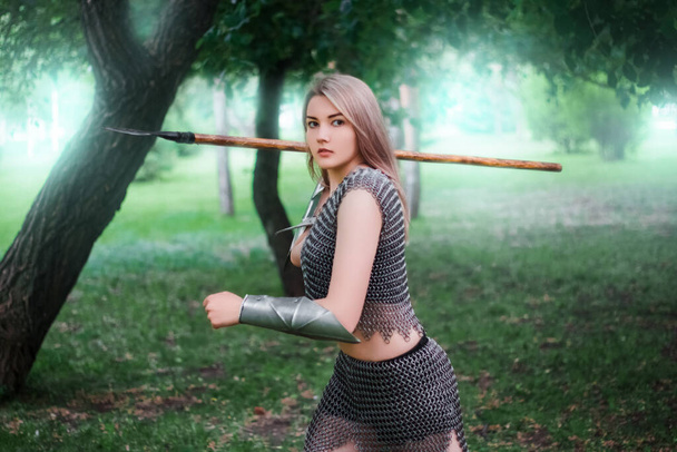 Portrait of a young medieval woman warrior, dressed in chain mail armor with a spear in her hands against the background of the dark forest. - 写真・画像
