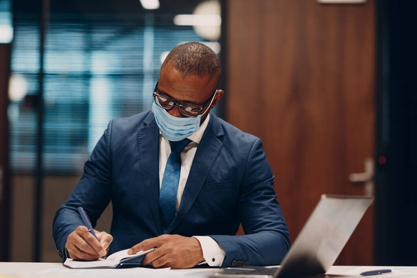 Portrait smiling african american businessman in medical face mask sit at table for meeting in office with notebook with pen and laptop. - Foto, Imagem