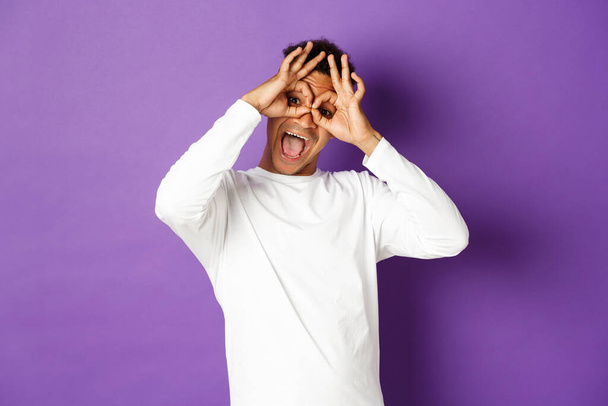 Image of funny and happy african-american man in white sweatshirt, grimacing and making faces, standing over purple background - Foto, immagini