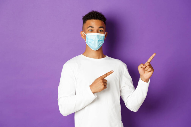 Fogalom a koronavírus, a karantén és az életmód. Jóképű afro-amerikai férfi orvosi maszkban, ujjal mutogat a jobb felső sarokban, logót mutat a fénymásolón, lila háttér - Fotó, kép