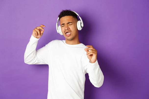 のんきなアフリカ系アメリカ人の男が踊り、無線のヘッドフォンで音楽を聞いて歌い、紫色の背景の上に立つ姿 - 写真・画像