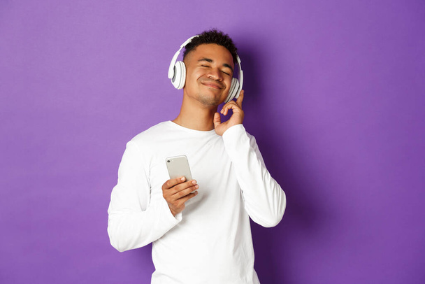 Imagen del hombre afroamericano despreocupado, disfrutando escuchando música en auriculares, sosteniendo el teléfono inteligente y sonriendo complacido, de pie sobre fondo púrpura - Foto, imagen
