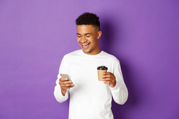 Happy african-american man drinking coffee, laughing and looking at smartphone, standing over purple background - Foto, Imagem