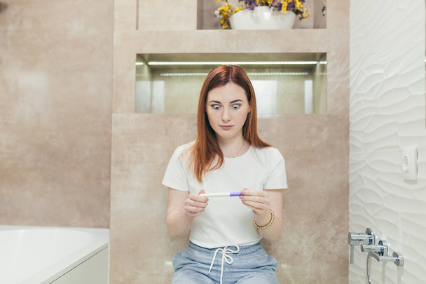 young frustrated woman looking at a quick positive or negative result pregnancy test sitting in bathroom by a unwanted. Female expectant mother sitting at home sad and disappointed - Фото, зображення