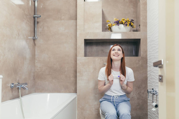 Happy caucasian young woman rejoices in positive pregnancy kit test for motherhood. Close up portrait of female at home checking result and emotions excited sitting sitting in the bathroom. Happiness - Foto, afbeelding