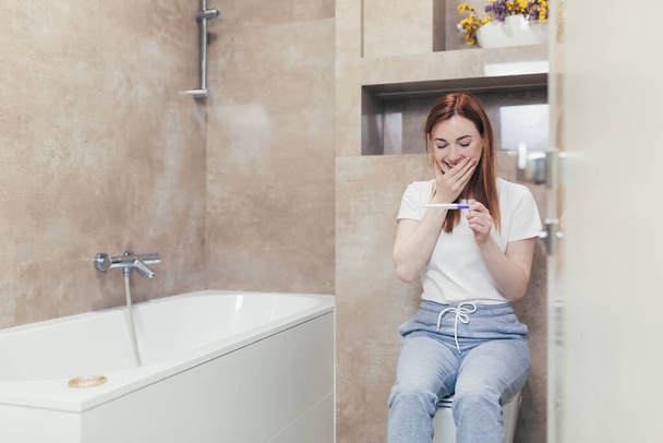 Happy caucasian young woman rejoices in positive pregnancy kit test for motherhood. Close up portrait of female at home checking result and emotions excited sitting sitting in the bathroom. Happiness - Foto, immagini