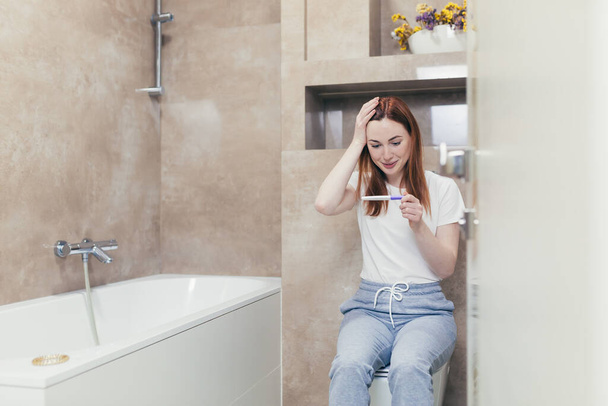 Happy caucasian young woman rejoices in positive pregnancy kit test for motherhood. Close up portrait of female at home checking result and emotions excited sitting sitting in the bathroom. Happiness - Foto, Imagem