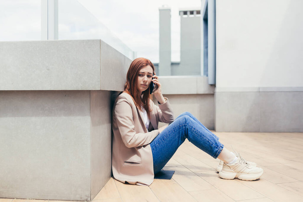 young frustrated woman sitting on sidewalk near a depressed business office center has lost job, financial problems. Female student girl stress at work or university college. Lady with grief outdoors - Фото, зображення