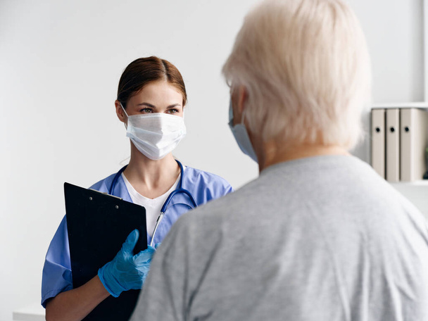 female nurse with documents in medical mask looking at elderly woman patient - Foto, Imagem