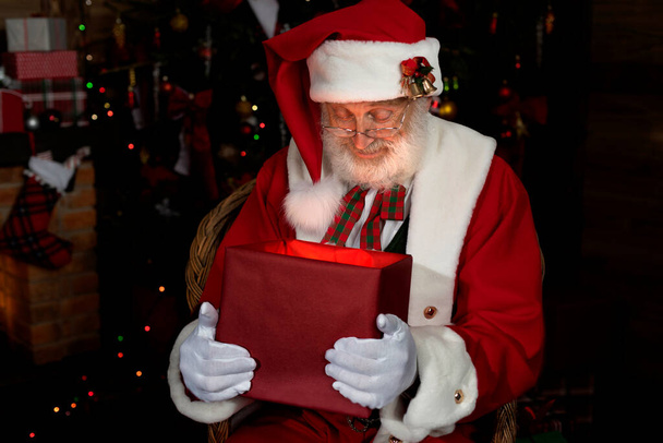 Санта-Клаус с подарочной коробкой возле камина и елки. Интерьер внутри деревянного дома, новогоднее бодрое настроение. Старший мужчина с настоящей белой бородой косплей Дед Мороз. - Фото, изображение