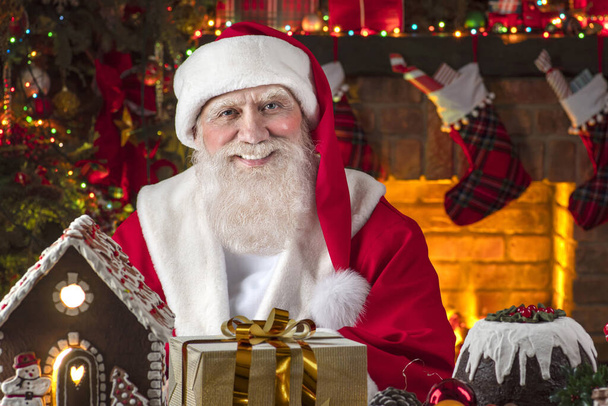 Noel Baba şöminenin ve Noel ağacının yanında. Ahşap evin içindeki şenlikli iç mekan, Noel 'in neşeli ruhu. Gerçek beyaz sakallı son sınıf öğrencisi Noel Baba kostümü giyiyor.. - Fotoğraf, Görsel