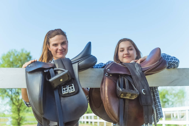Dos chicas jóvenes en el rancho de caballos sillas de cuero colgando de la valla de madera  - Foto, Imagen