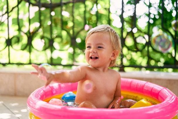 Piccolo bambino sorridente siede in una piccola piscina gonfiabile e cattura bolle di sapone - Foto, immagini