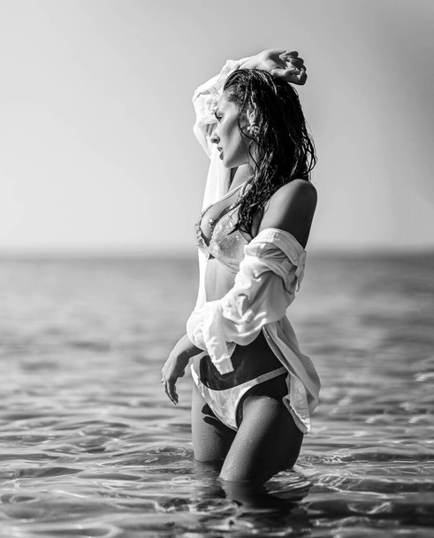 Young woman in white bikini and shirt stands in waist-deep sea, looking into the distance. Black and white. Summer vibes - Фото, изображение