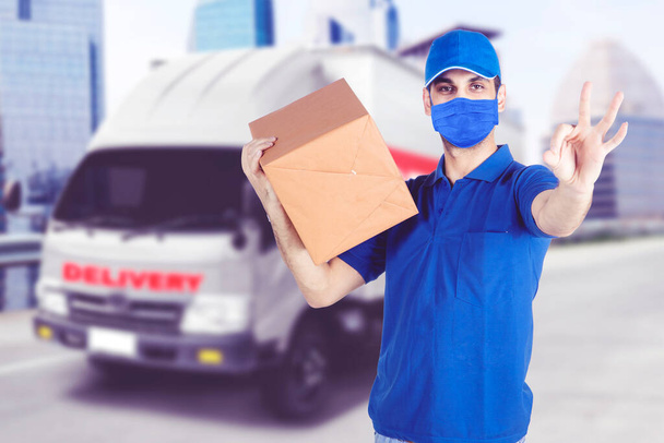 Male courier showing OK sign and standing near the delivery truck while wearing face mask during new normal lifestyle - Photo, Image